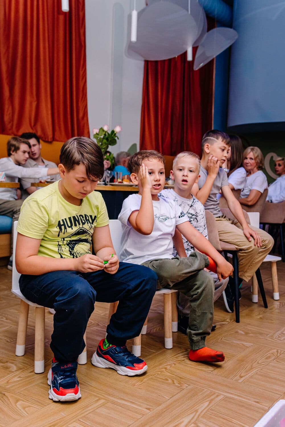 Мальчики сидят рядком на детских стульчиках и смотрят шоу, за ними столы с гостями