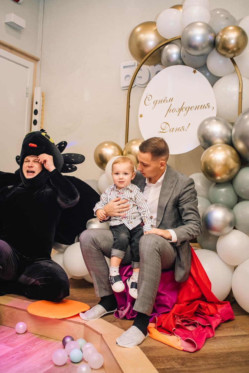 Папа держит малыша-именинника у фотозоны с белыми, серыми и золотыми шарами, рядом артистка в чёрном костюме Беззубика