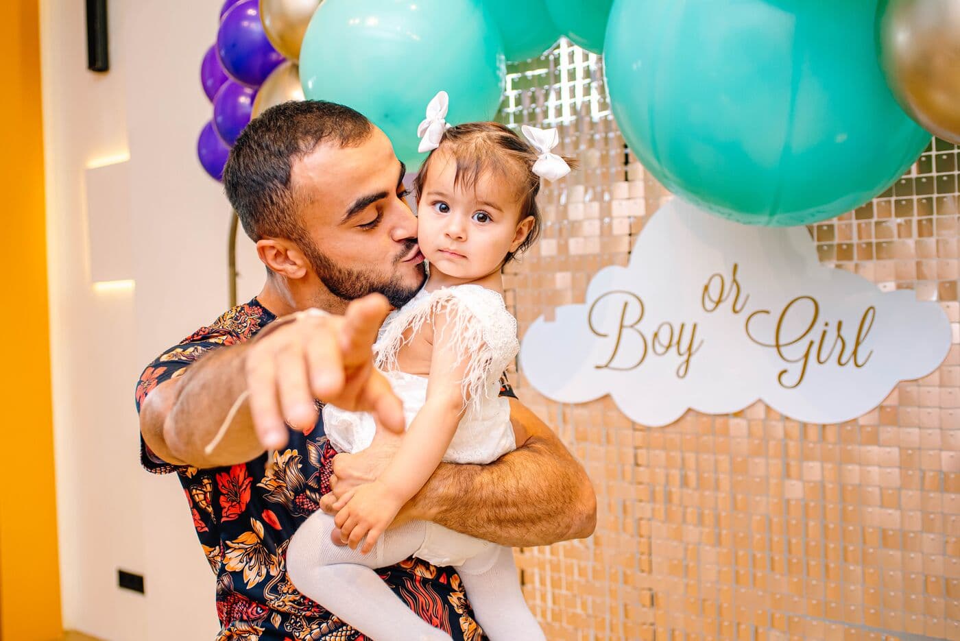 Папа целует дочку на руках у фотозоны с надписью «Boy or Girl» и бирюзовыми шарами