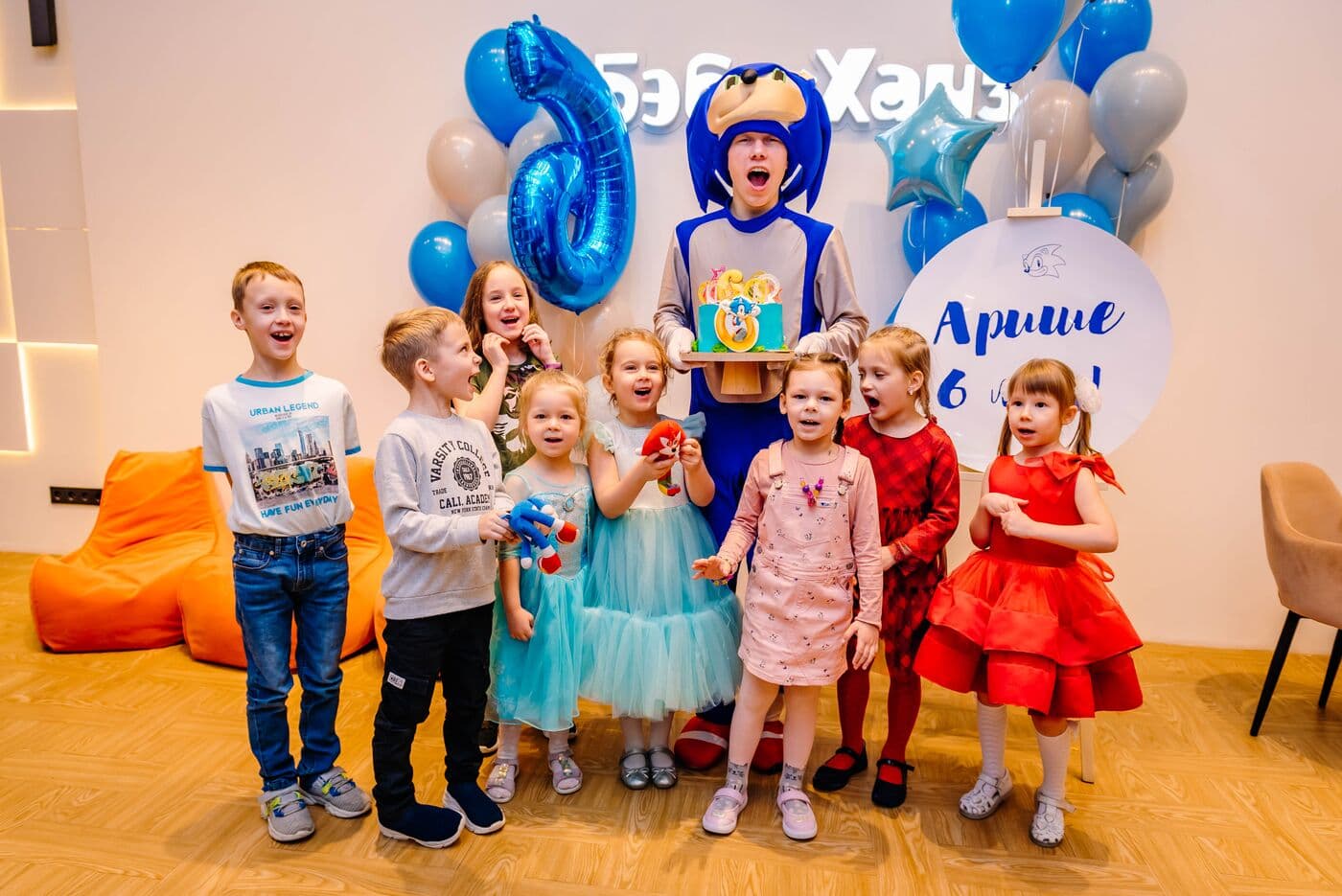 Дети с восторгом позируют у фотозоны с синими шарами, синей цифрой 6 и надписью «Бэби Хауз», в центре артист в костюме Соника с тортом