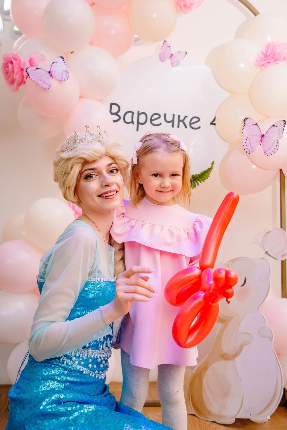 Артистка в образе Эльзы с короной обнимает девочку в розовом платье с шариком-собачкой